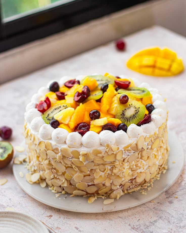 A fresh fruit cake topped with sliced kiwi, cherries, mango chunks, and grapes, surrounded by white whipped cream and coated with sliced almonds on the sides.